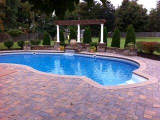Swimming pool with blue water and stone patio, with a pergola and landscaping in the background.