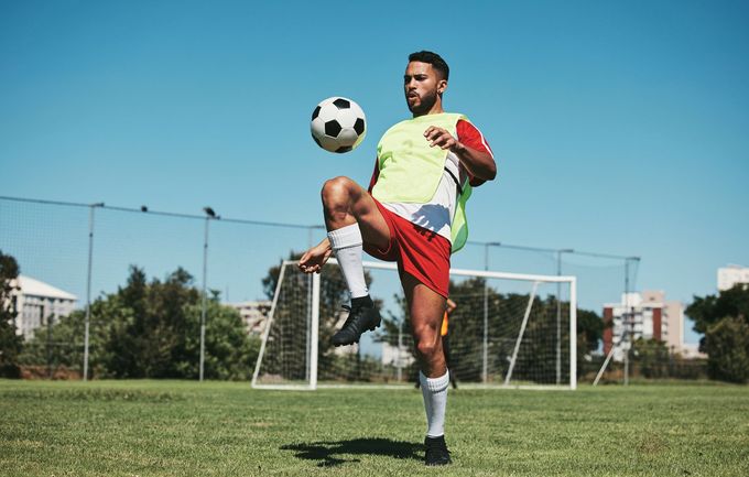 Soccer player juggling a ball on a grassy field in front of a goal under a clear blue sky