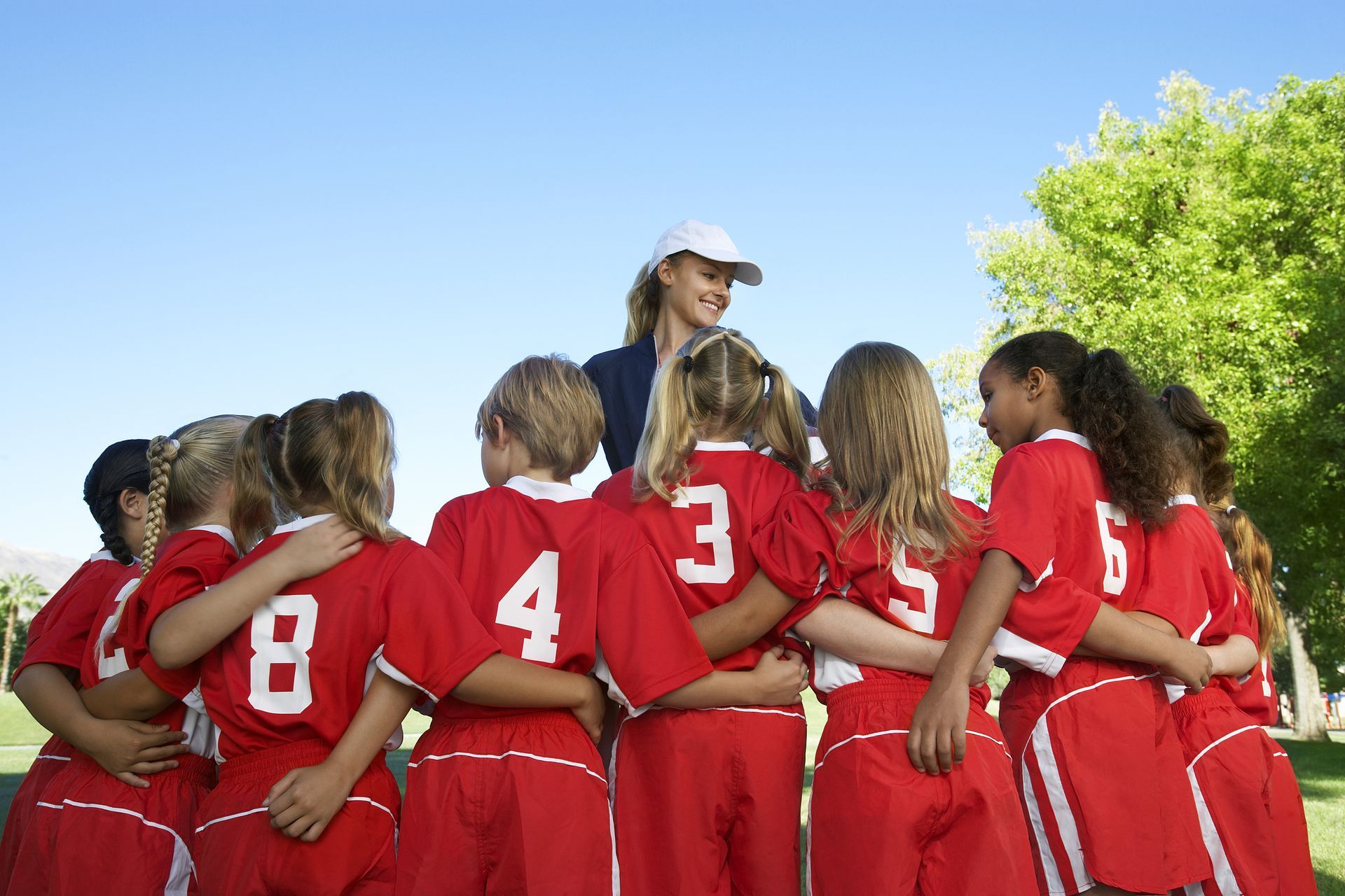 Youth soccer team in red jerseys huddles around coach on a sunny field.