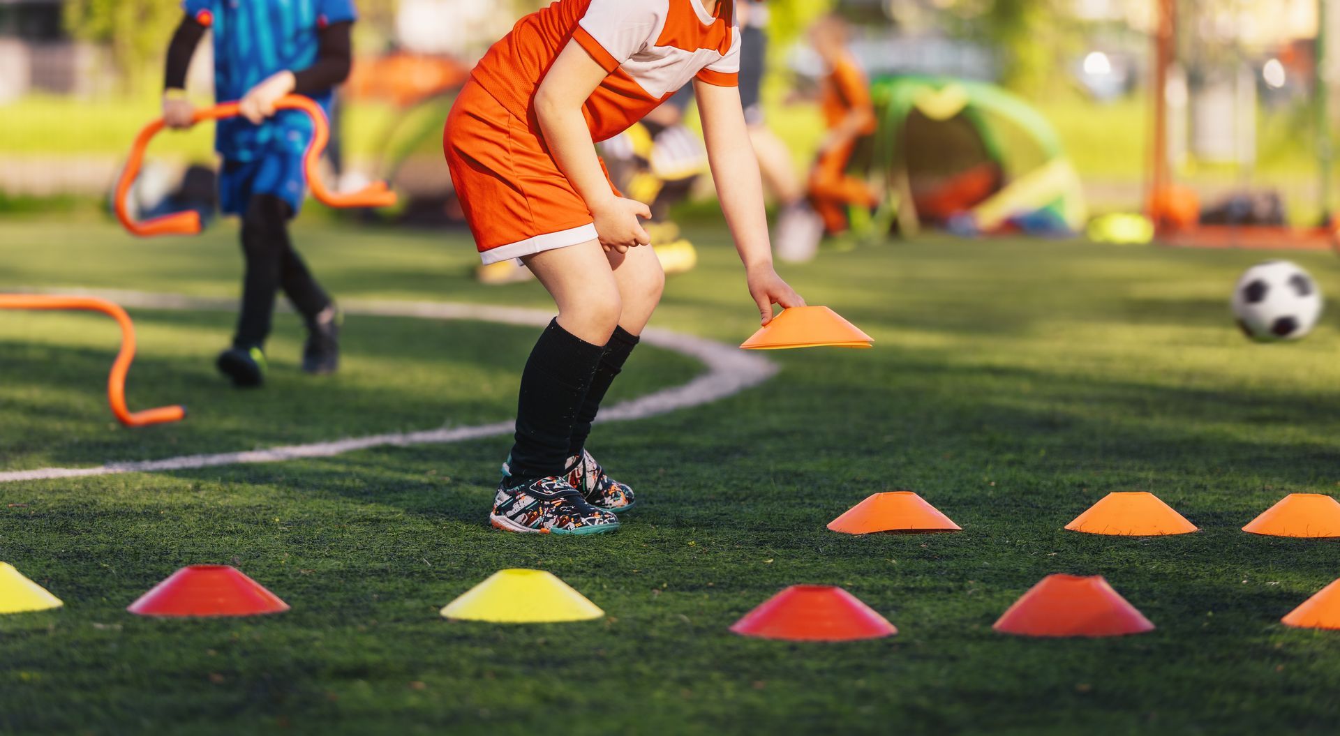 Children practicing soccer drills with orange cones on a grassy field, with one player in orange uniform