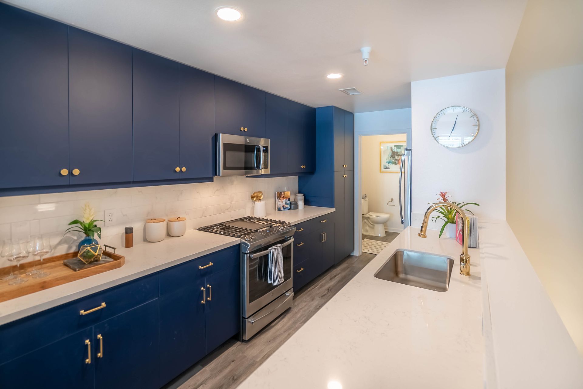 A kitchen with blue cabinets , stainless steel appliances , a sink , and a clock on the wall.