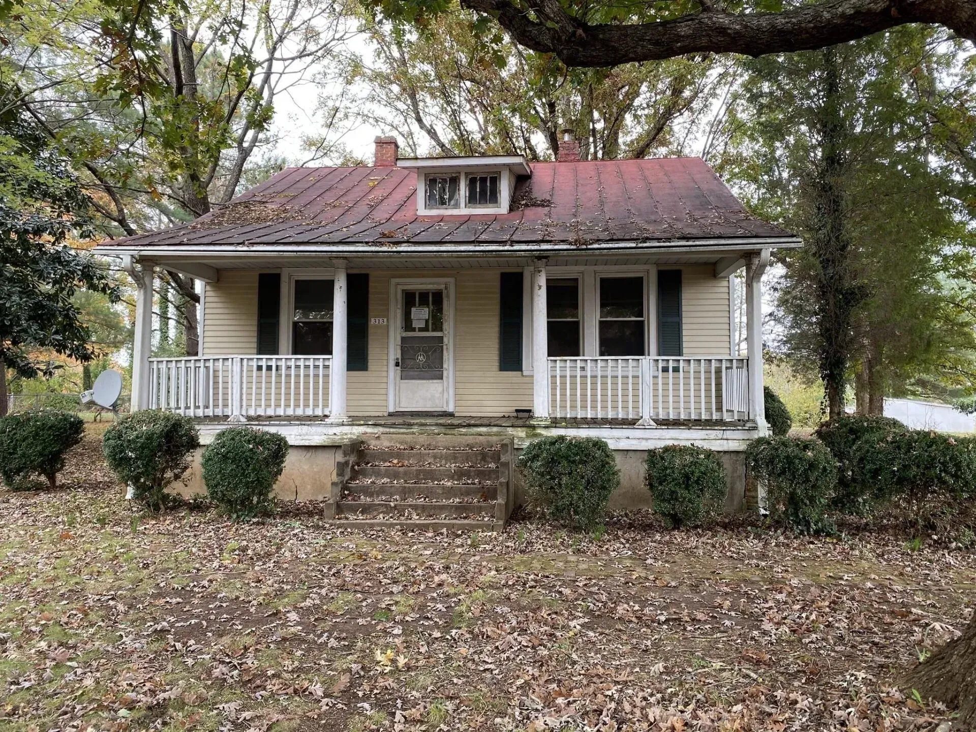 A small house with a porch and a red roof is surrounded by trees and leaves.