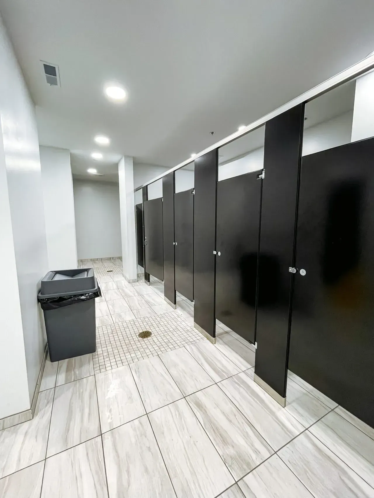 A public restroom with black stalls and a trash can.
