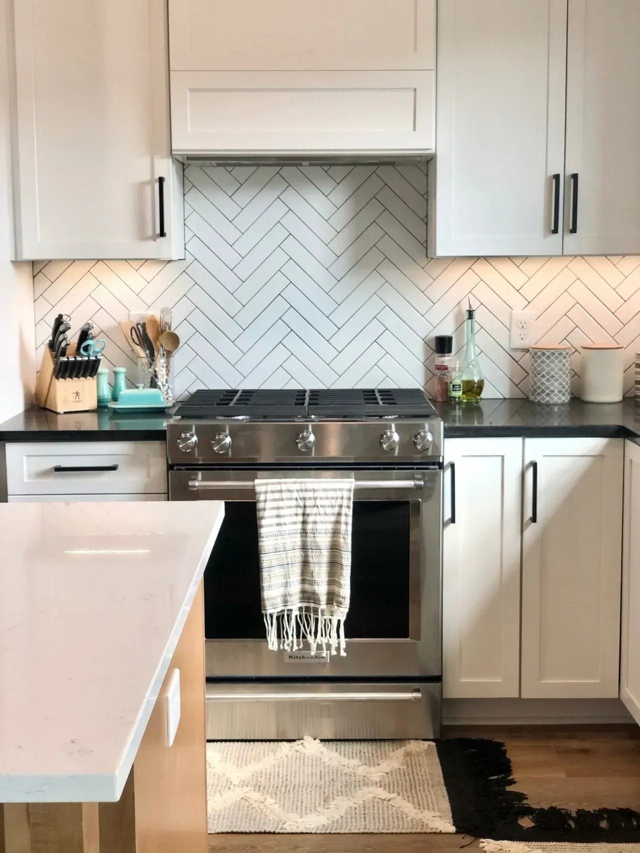 A kitchen with stainless steel appliances , white cabinets , and a herringbone tile wall.