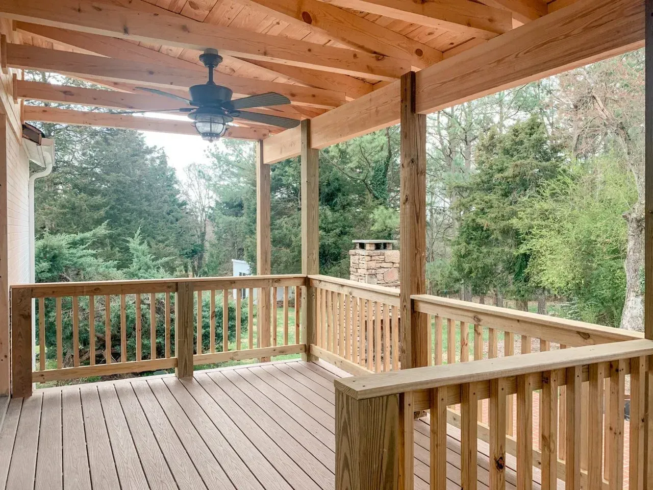 A wooden deck with a ceiling fan and trees in the background.