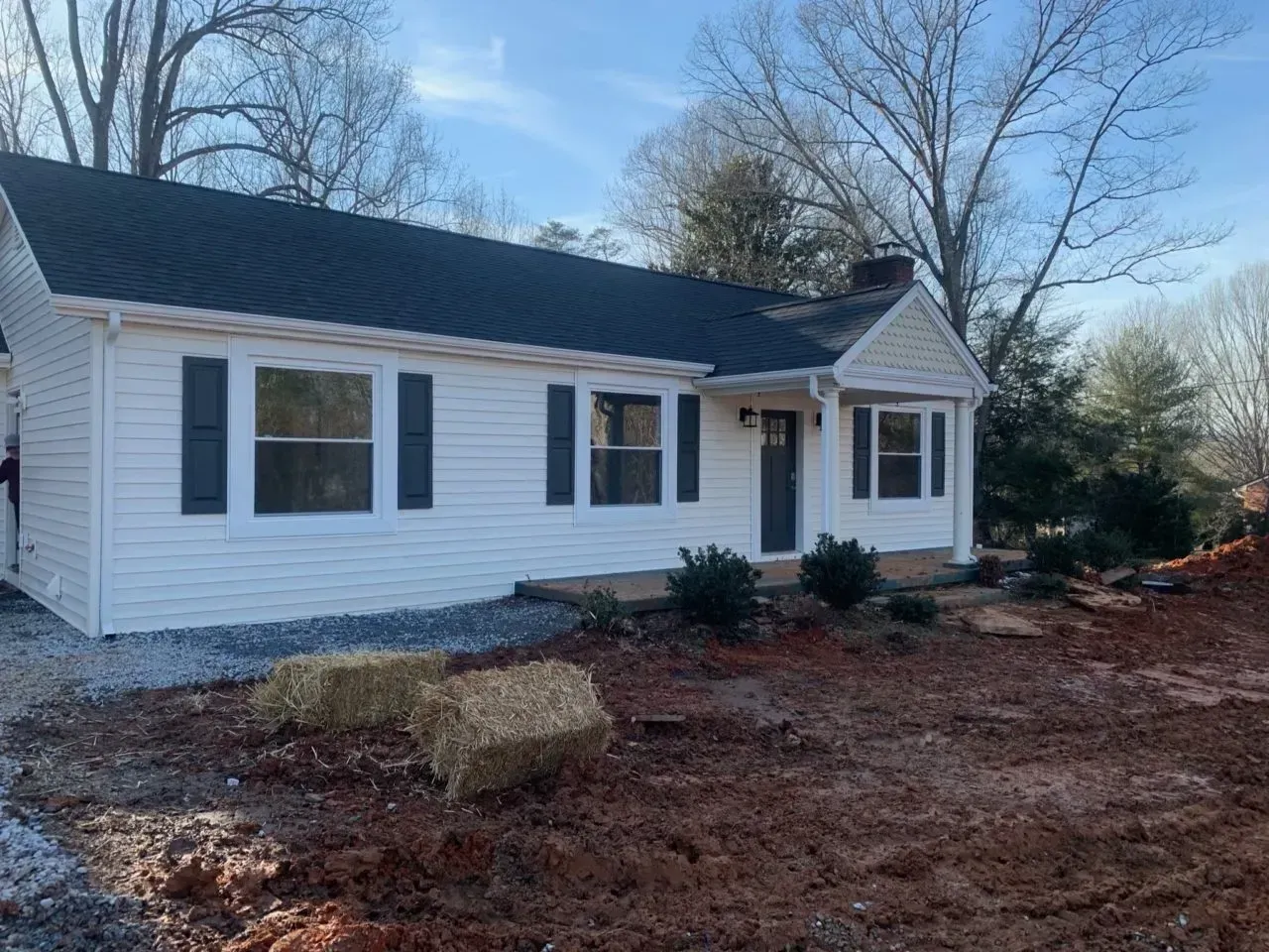A white house with black shutters is sitting in the middle of a dirt field.