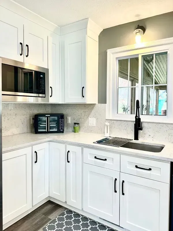 A kitchen with white cabinets , a sink , a microwave , and a window.