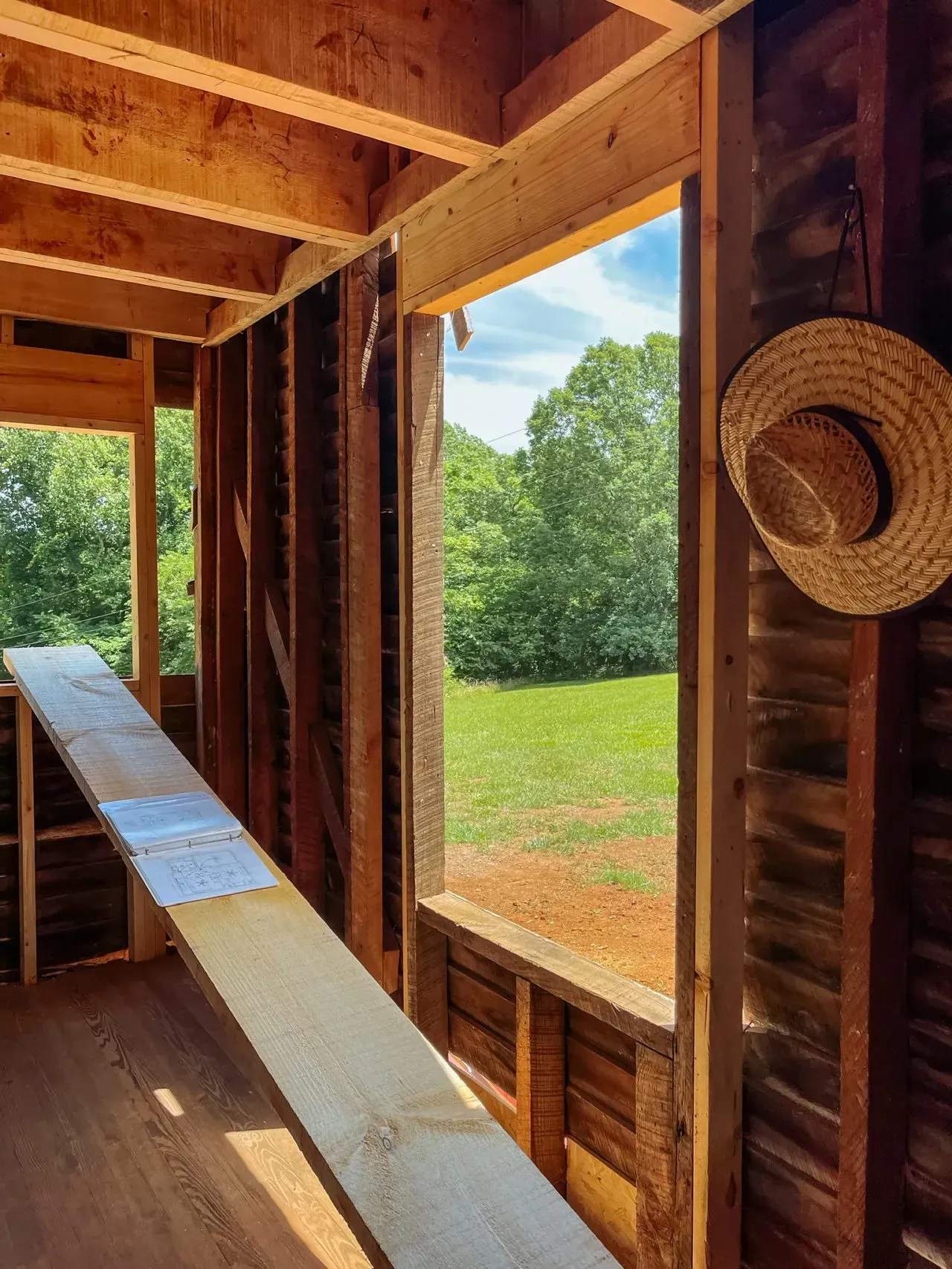 A straw hat is hanging on a wooden wall next to a window.
