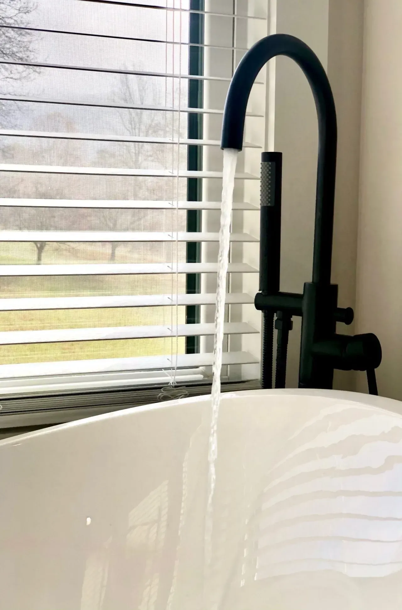 A bathtub with water running from a black faucet in front of a window with blinds.