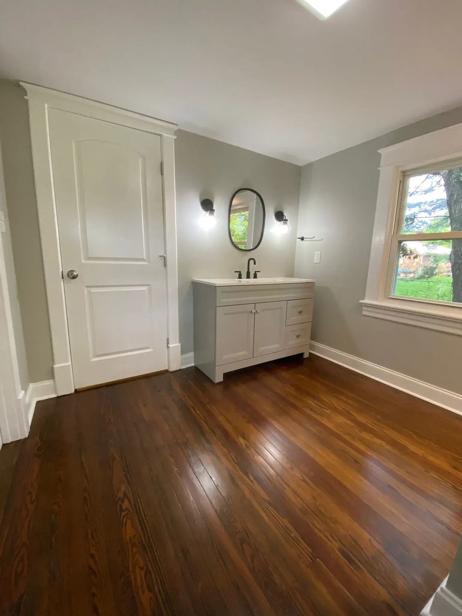 A bathroom with hardwood floors , a sink , a mirror and a window.