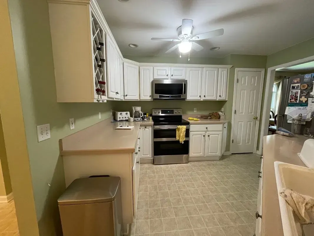A kitchen with white cabinets , stainless steel appliances , a trash can and a ceiling fan.
