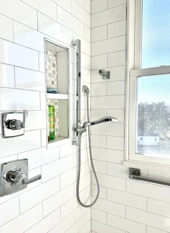 A bathroom with white tiles , a shower head , and a window.