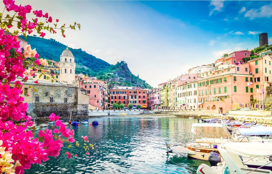 Colorful waterfront village with boats, pastel buildings, mountains, and bright pink flowers