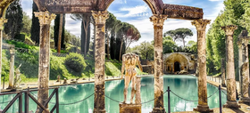 Ancient stone ruins around a turquoise pool with a small fountain and distant waterfall under a blue sky