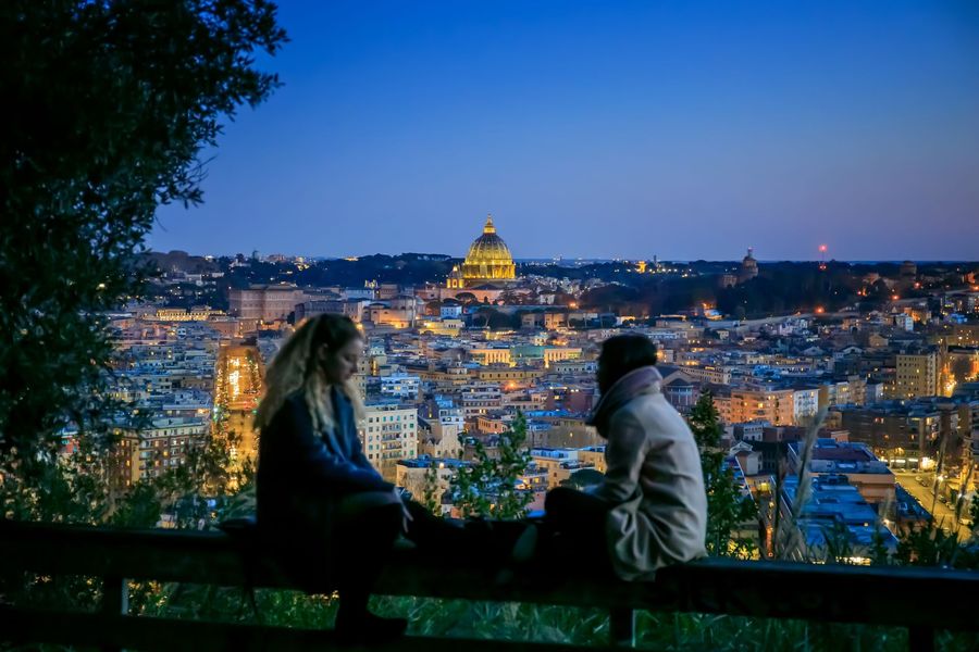 Two people sit on a bench overlooking a lit city at dusk, with a golden domed landmark in the distance.