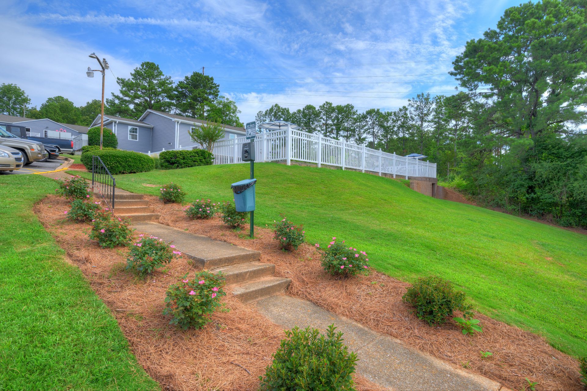 Concrete steps leading up a grassy hill towards a white fence, surrounded by a mulched garden bed with small bushes.