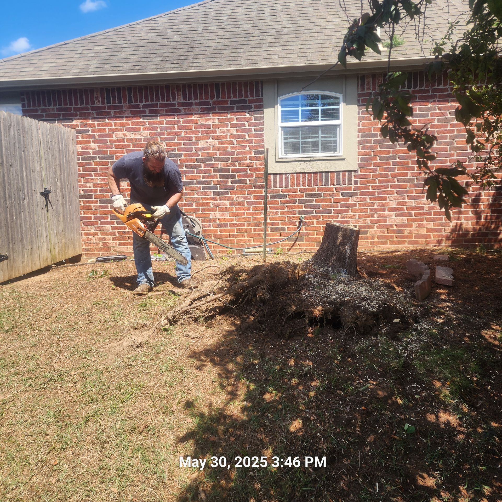 A man is cutting a tree stump in front of a brick house