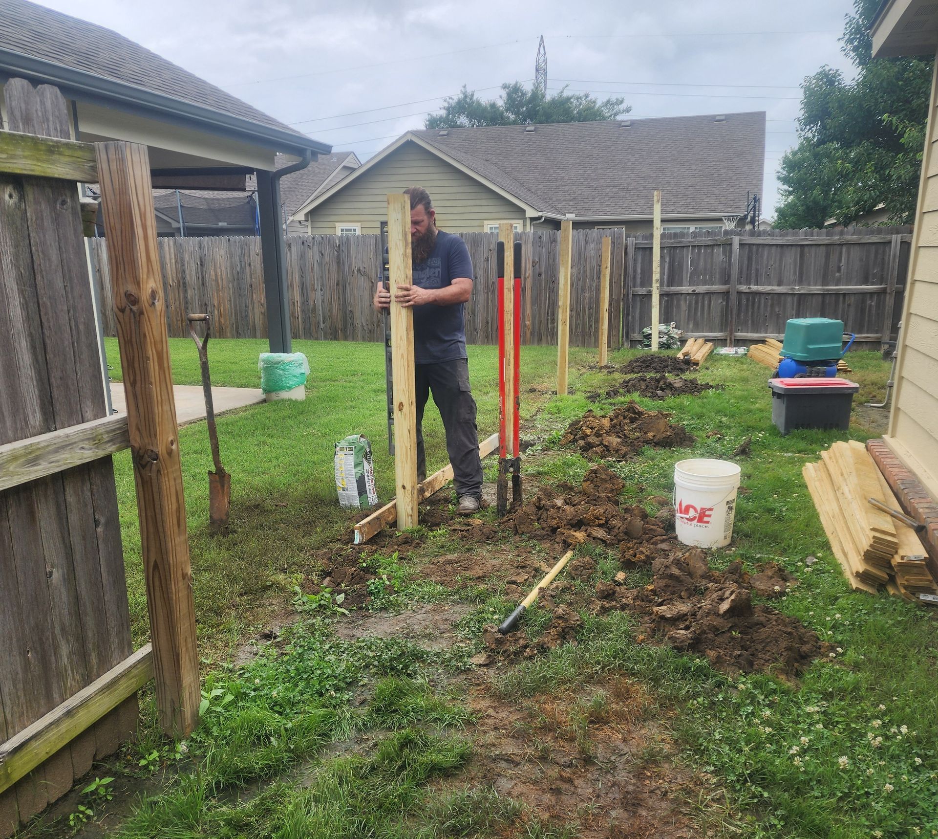 A man is standing in the dirt in a backyard holding a wooden post.