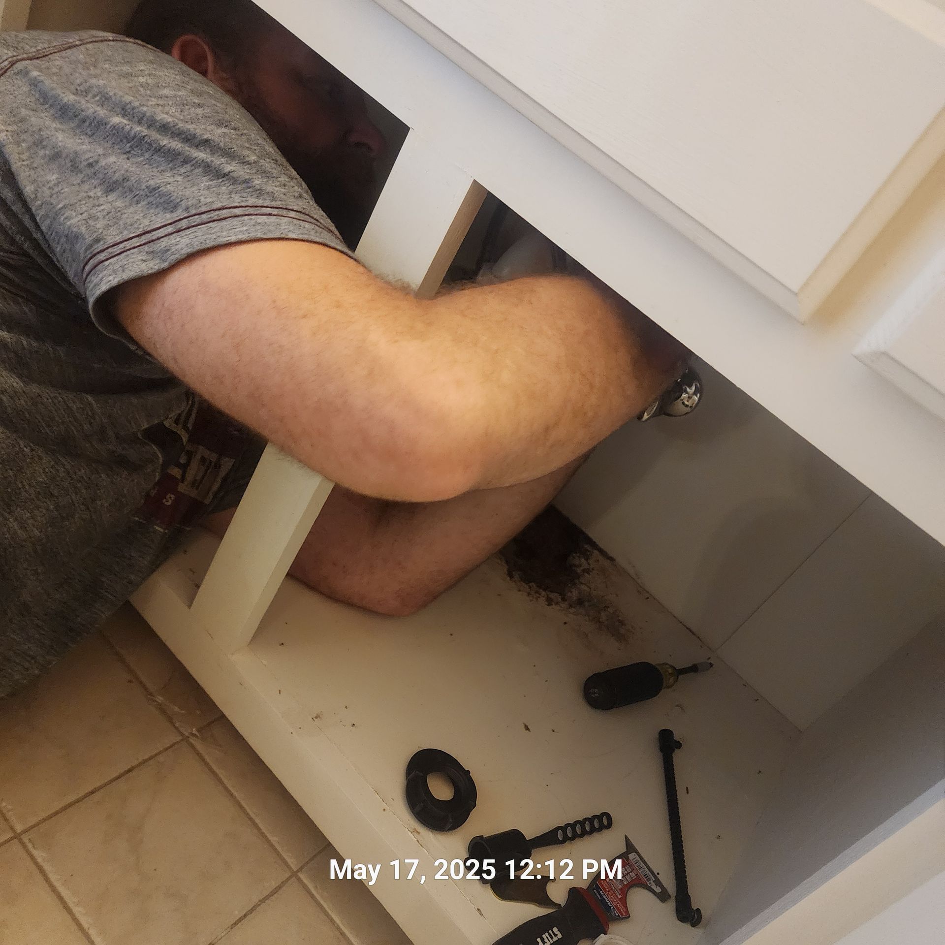 A man is working under a sink on may 17th at 12:12 pm