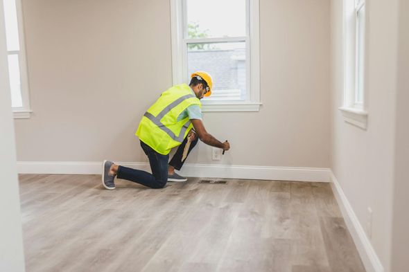 Man in safety vest and hardhat kneels, measuring a wall's baseboard in a room with light-colored walls and floors.
