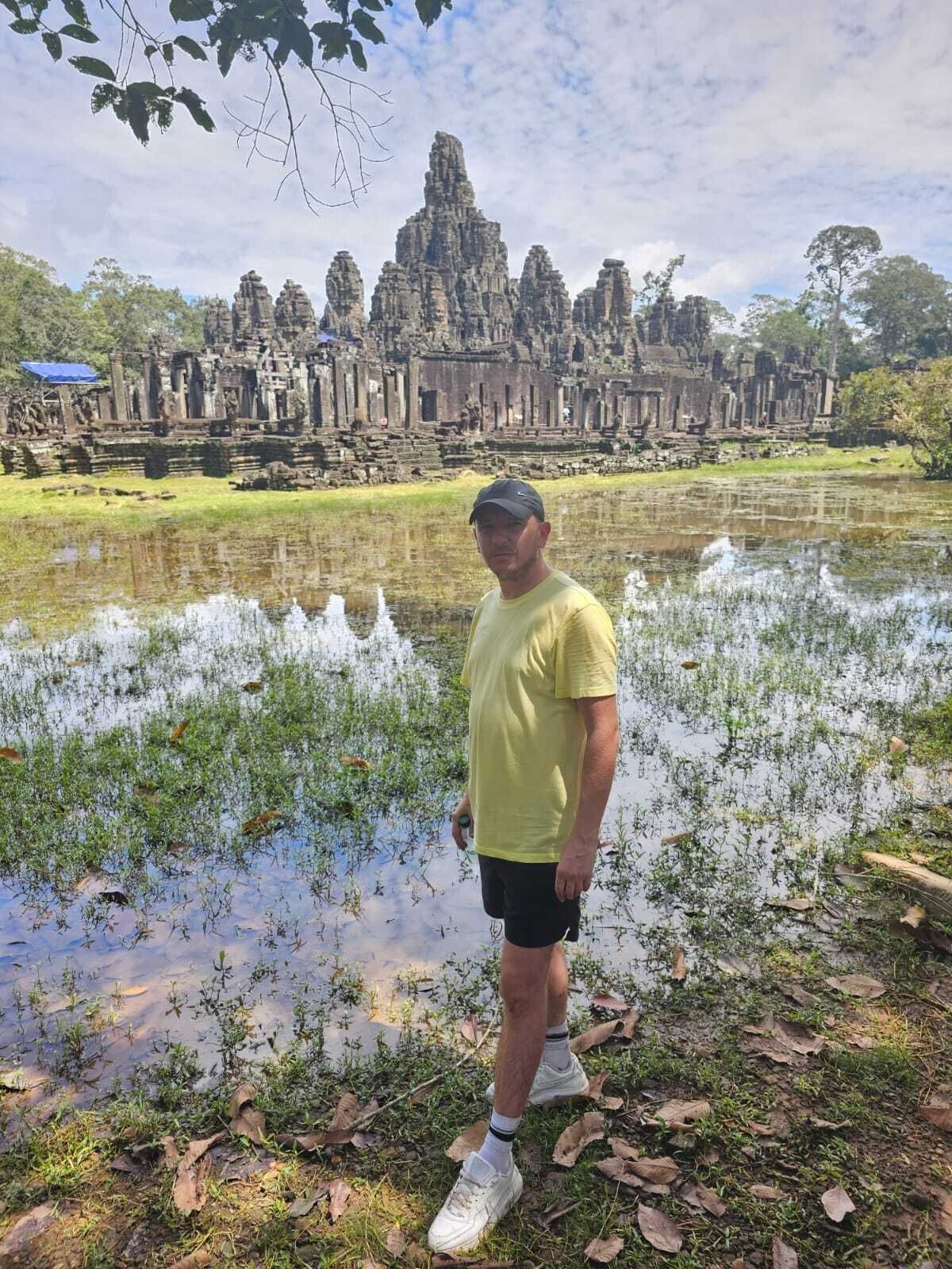 Man in yellow shirt poses near a pond in front of Angkor Wat temple.