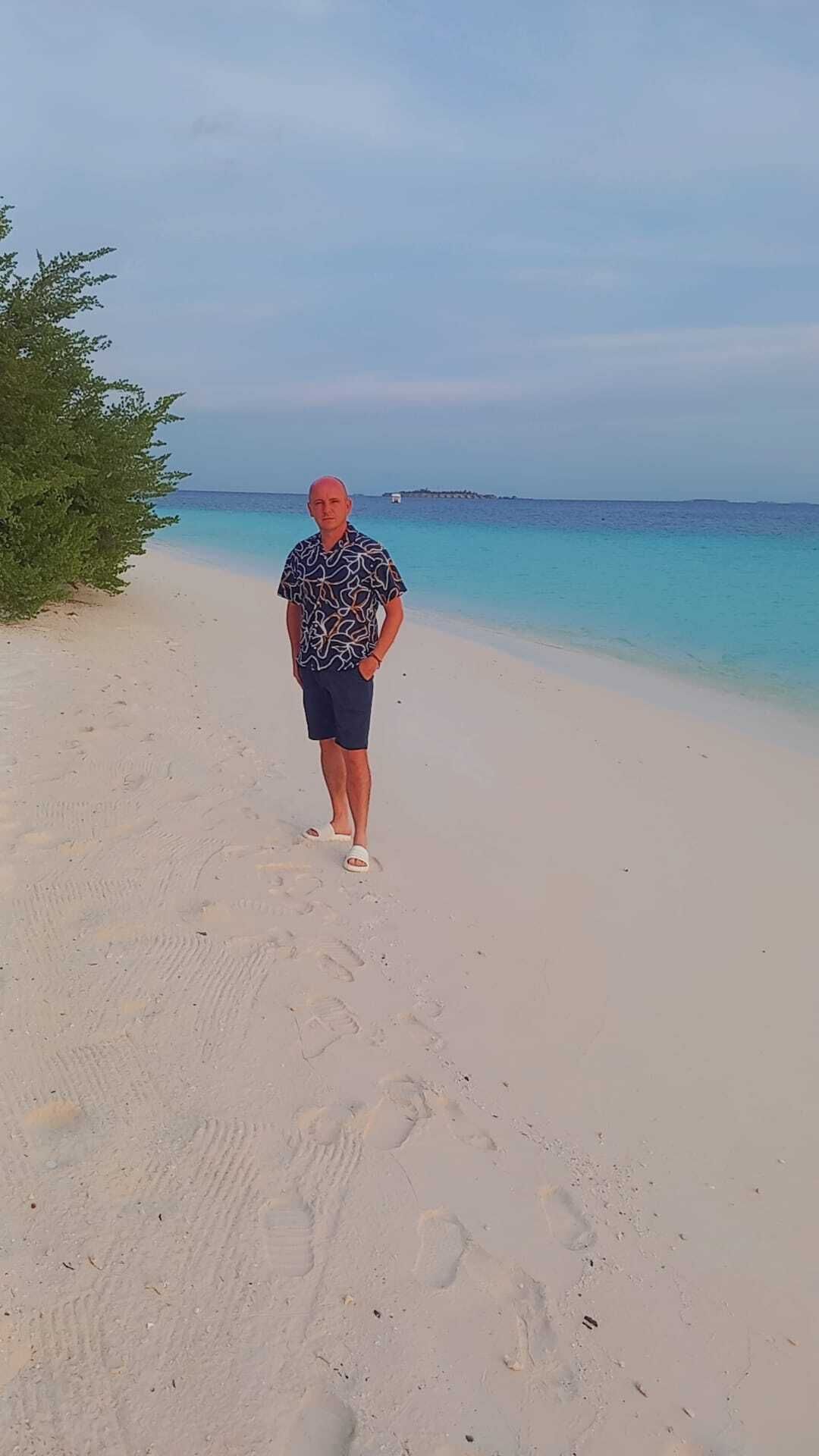 Man on white sand beach, wearing blue shorts, floral shirt, and red cap, with turquoise water.