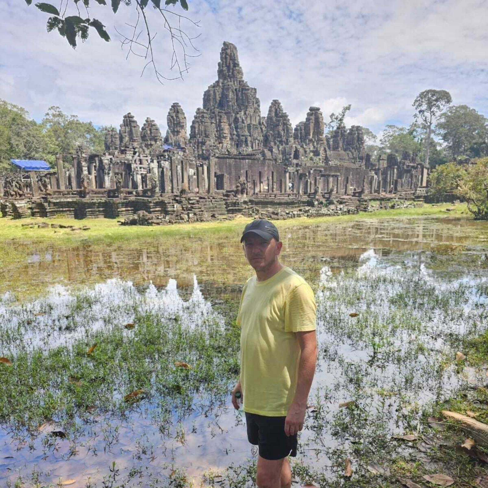 Man in yellow shirt and cap stands before ancient temple, reflecting in water.