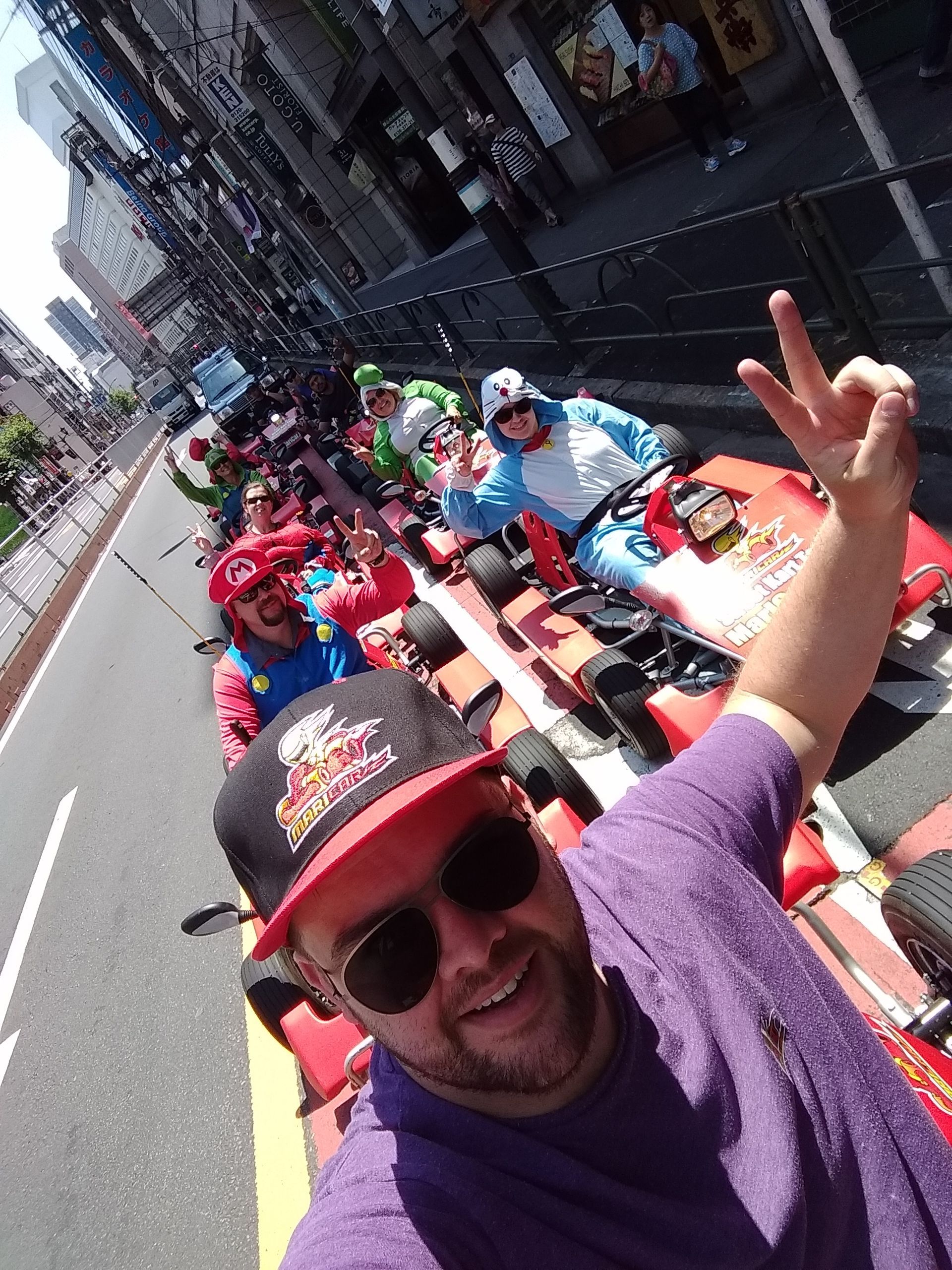 Man in purple shirt on go-kart, making peace sign. Others in costumes and karts parade on city street.