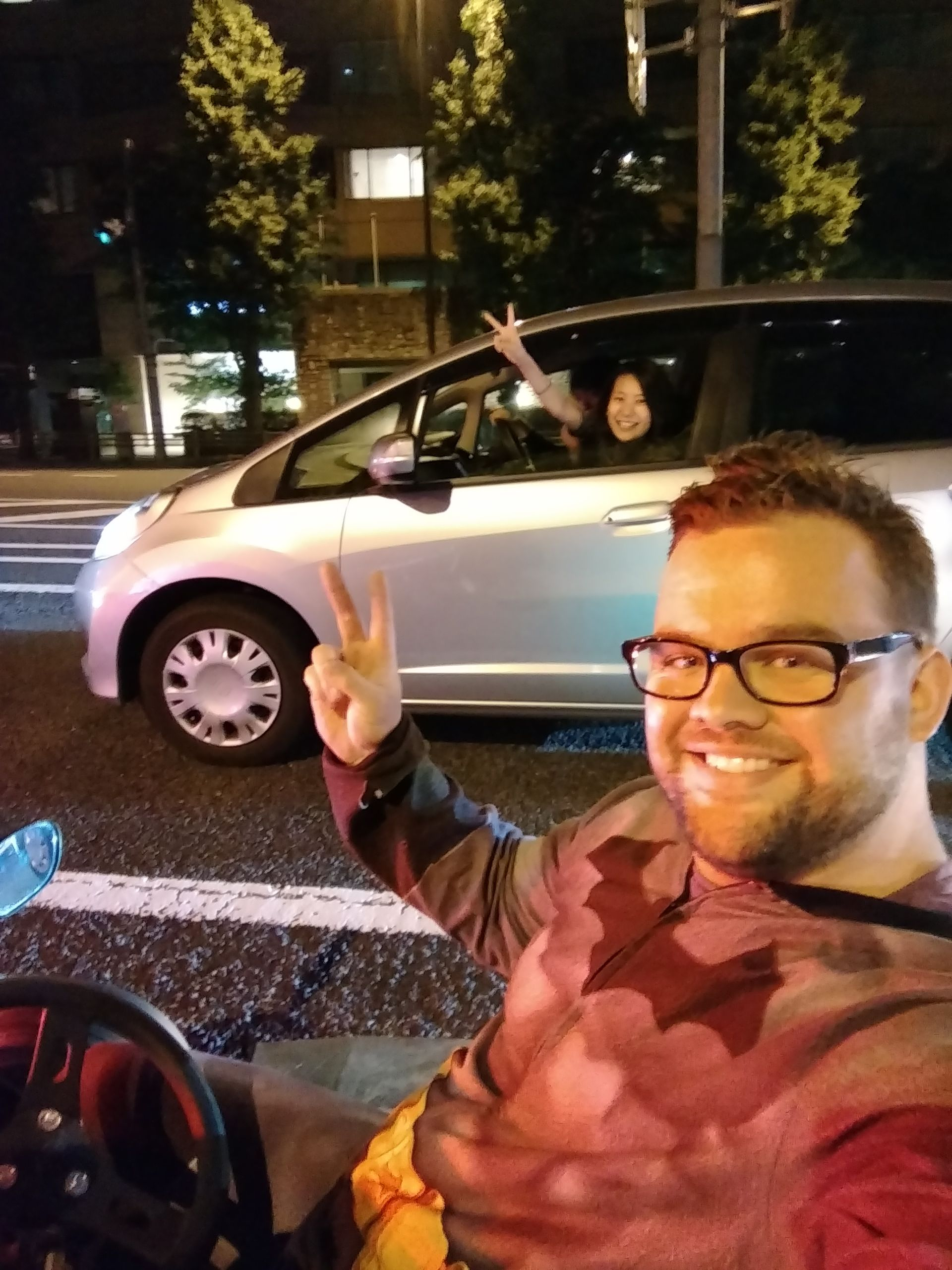 Man in costume smiles, giving peace sign as woman in car waves. Nighttime, city street.