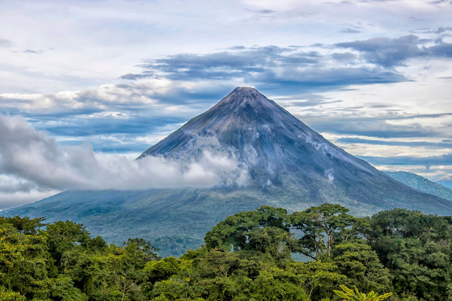 Arenal volcano, costa rica covered in clouds is surrounded by trees and mountains.