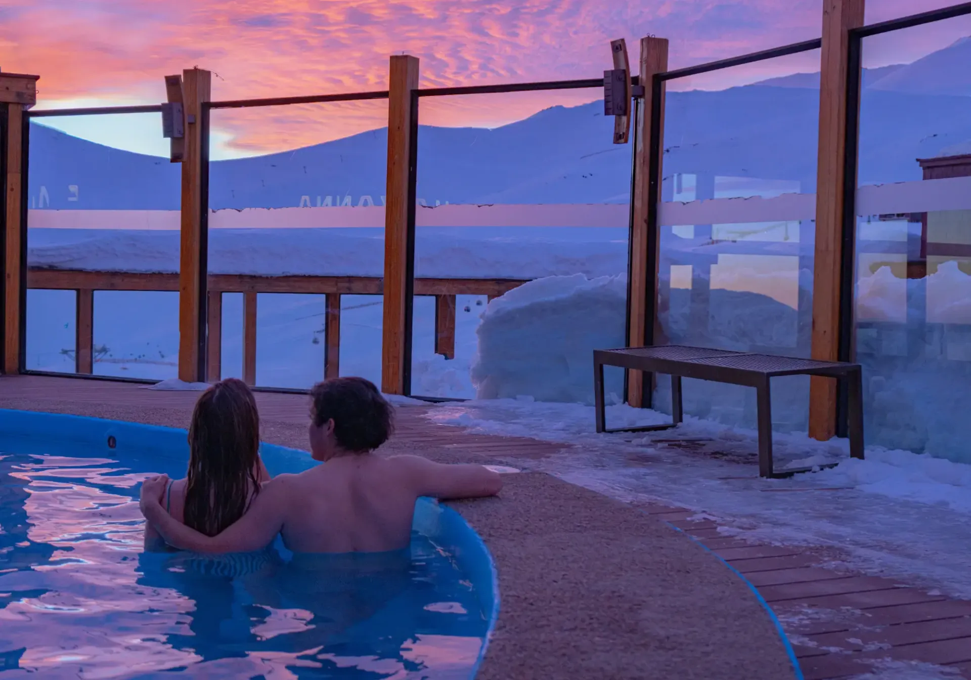 Couple relaxing in a hot tub at sunset beside snowy mountains and a purple sky.