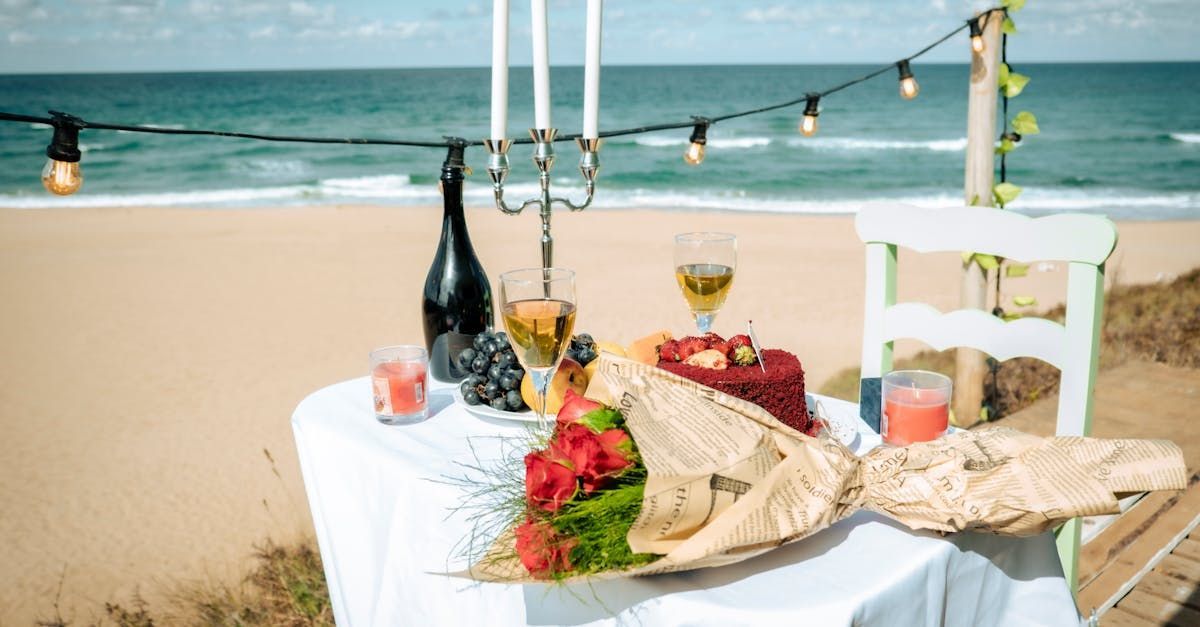 A table with a cake , wine , candles and flowers on it on the beach.
