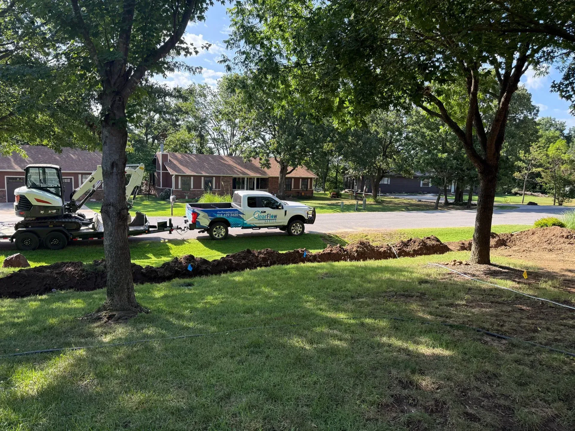 A white truck and excavator digging a trench in a grassy front yard, near a residential street.