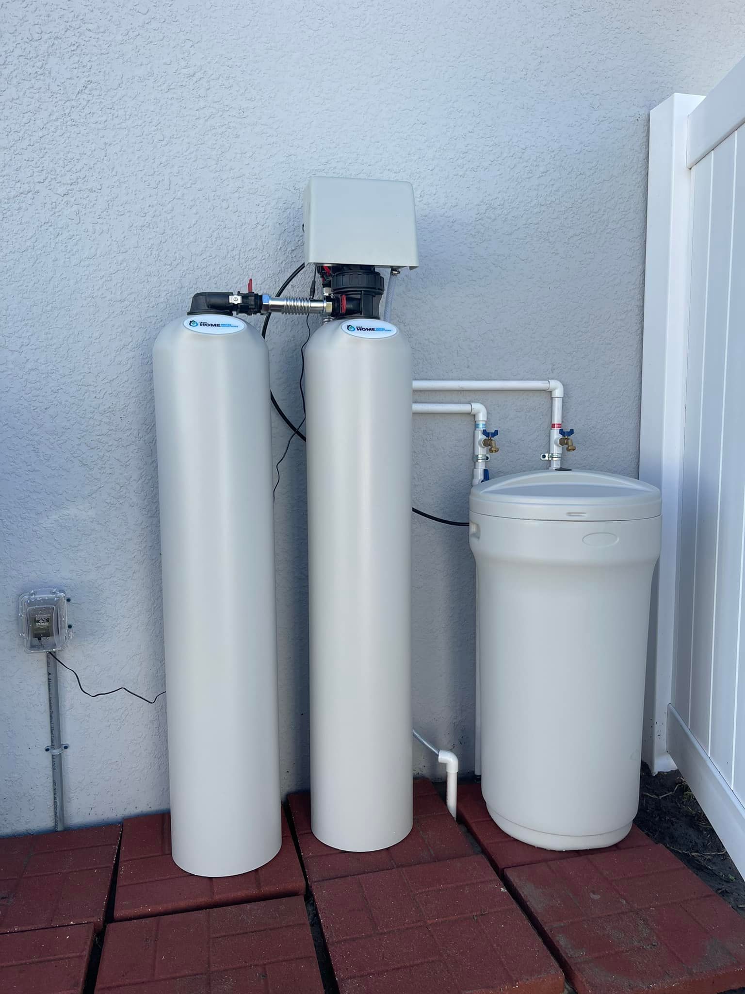 Water softener system against a stucco wall, with three white tanks and plumbing.