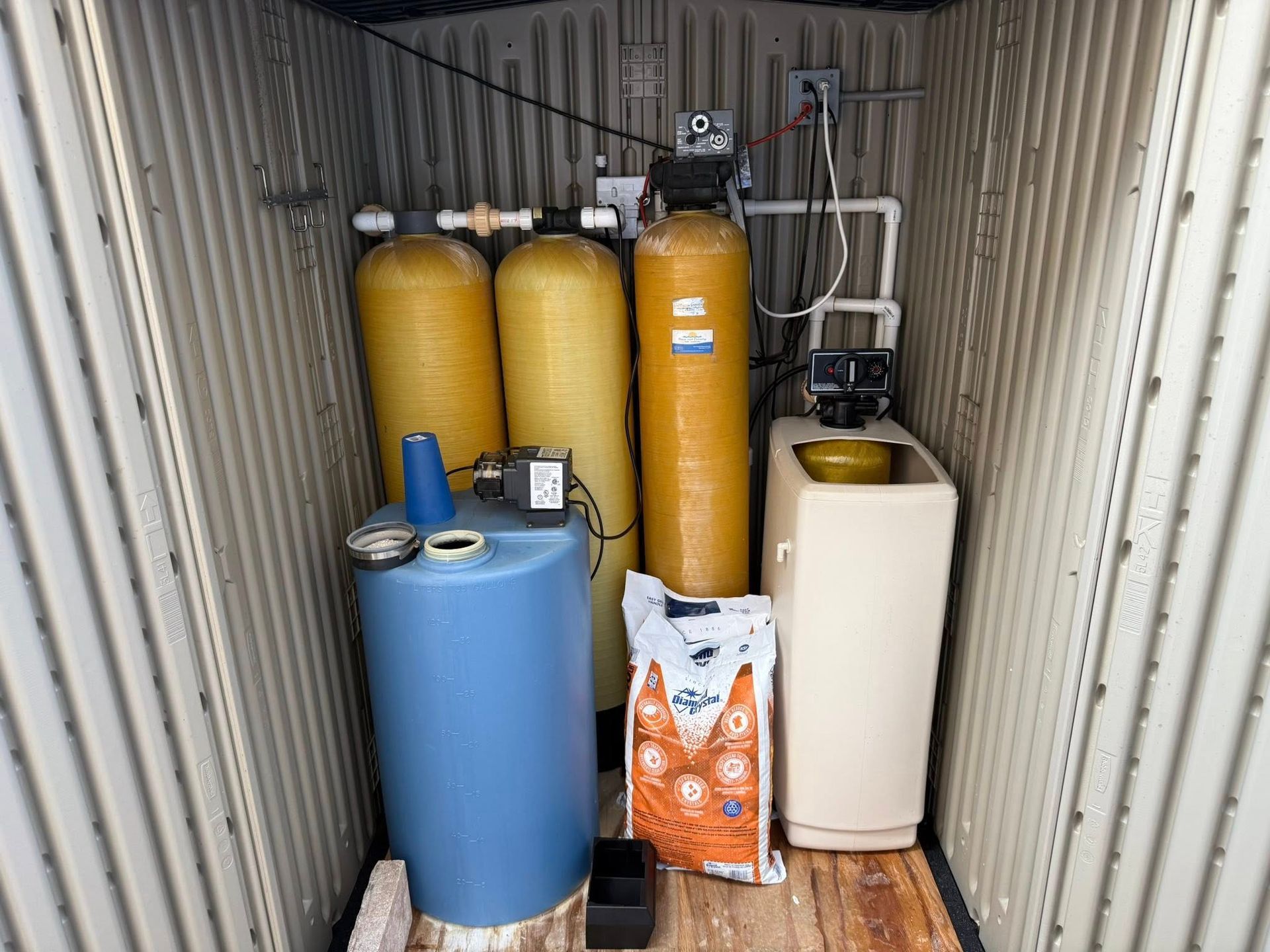 Water filtration system inside a metal enclosure, featuring yellow and blue tanks and a bag of pellets.