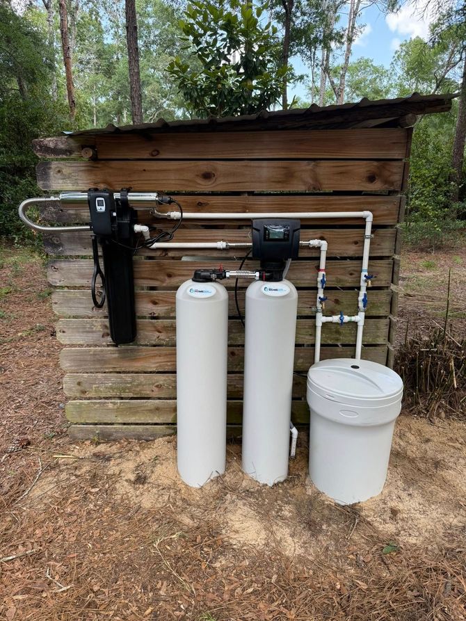Water filtration system outdoors, housed in a wooden shed. Includes filter, softener tanks, and salt container.