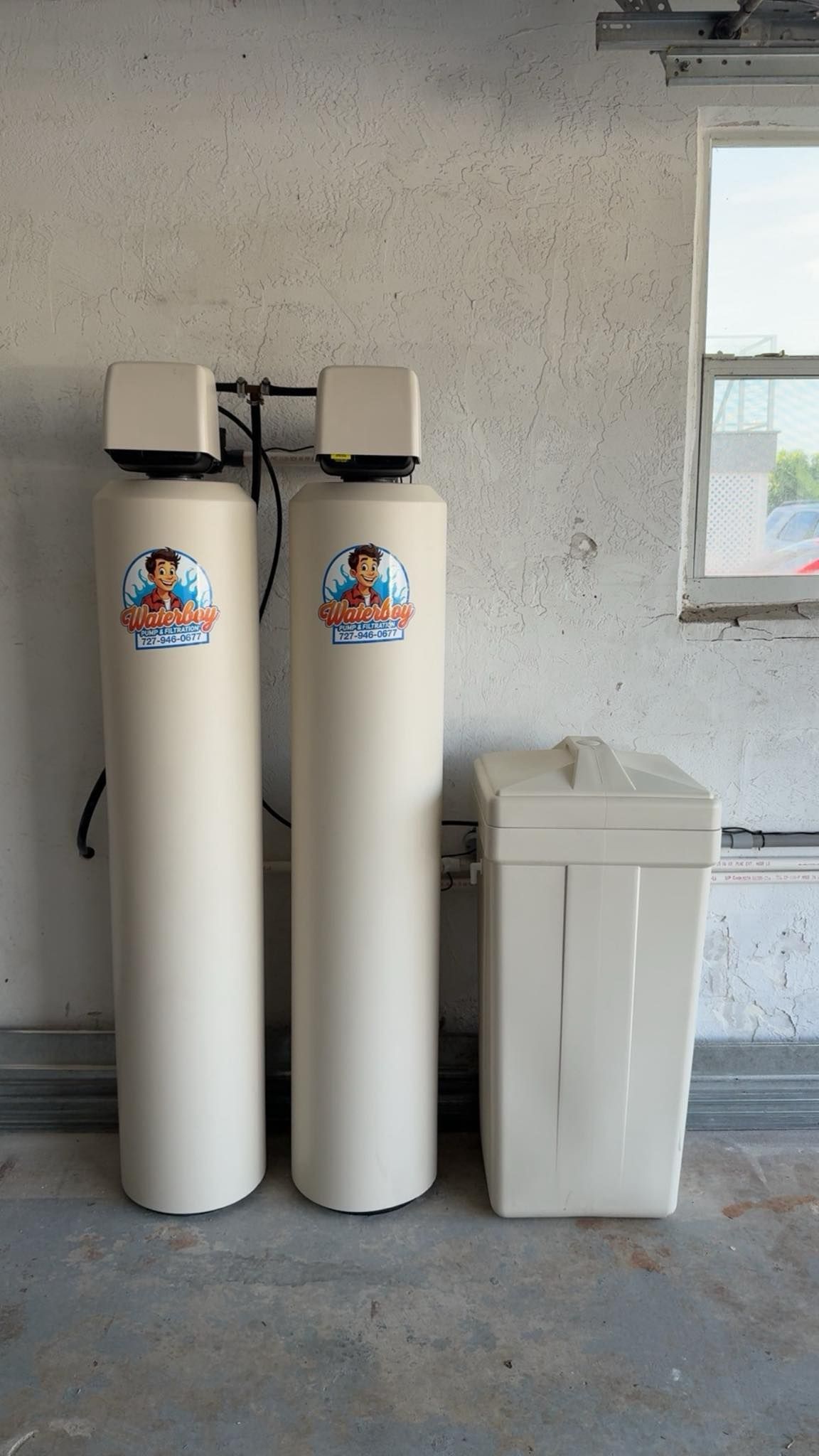 Water filtration system in a garage. Two tall beige tanks and a white salt tank against a concrete wall.