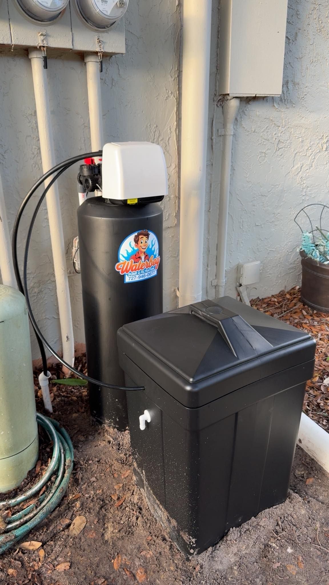 Water softener system outdoors, black tank with control head and salt container, beside stucco wall.