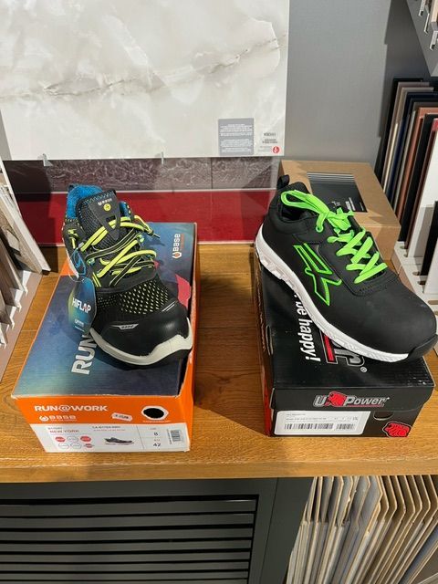 Due paia di scarpe antinfortunistiche in scatole, nere con dettagli verde neon, su uno scaffale di legno.