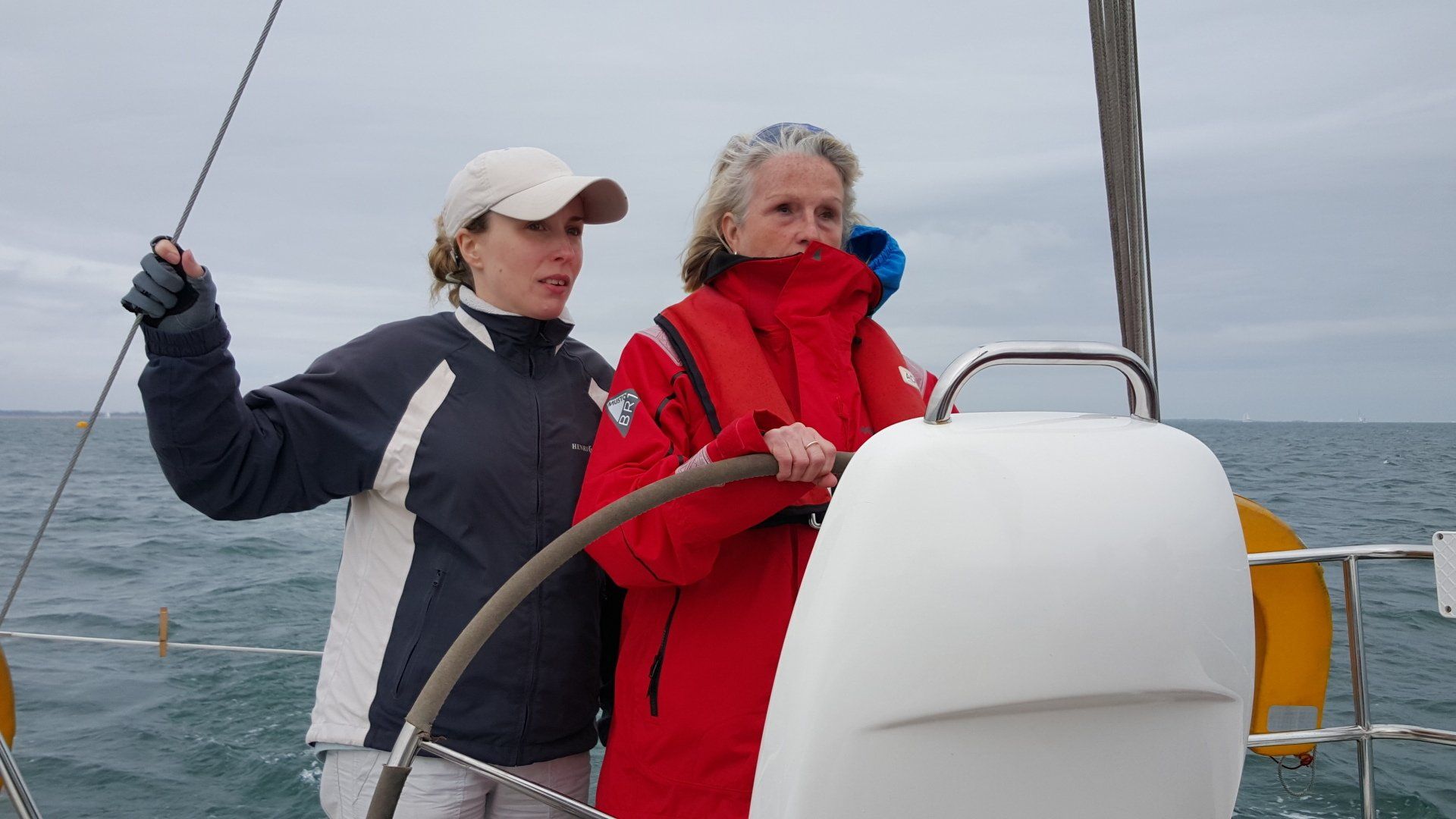 An image of a V.I. sailor at the helm of an underway yacht, being guided by a sighted skipper. They are in wet weather gear, and the wind is strong.