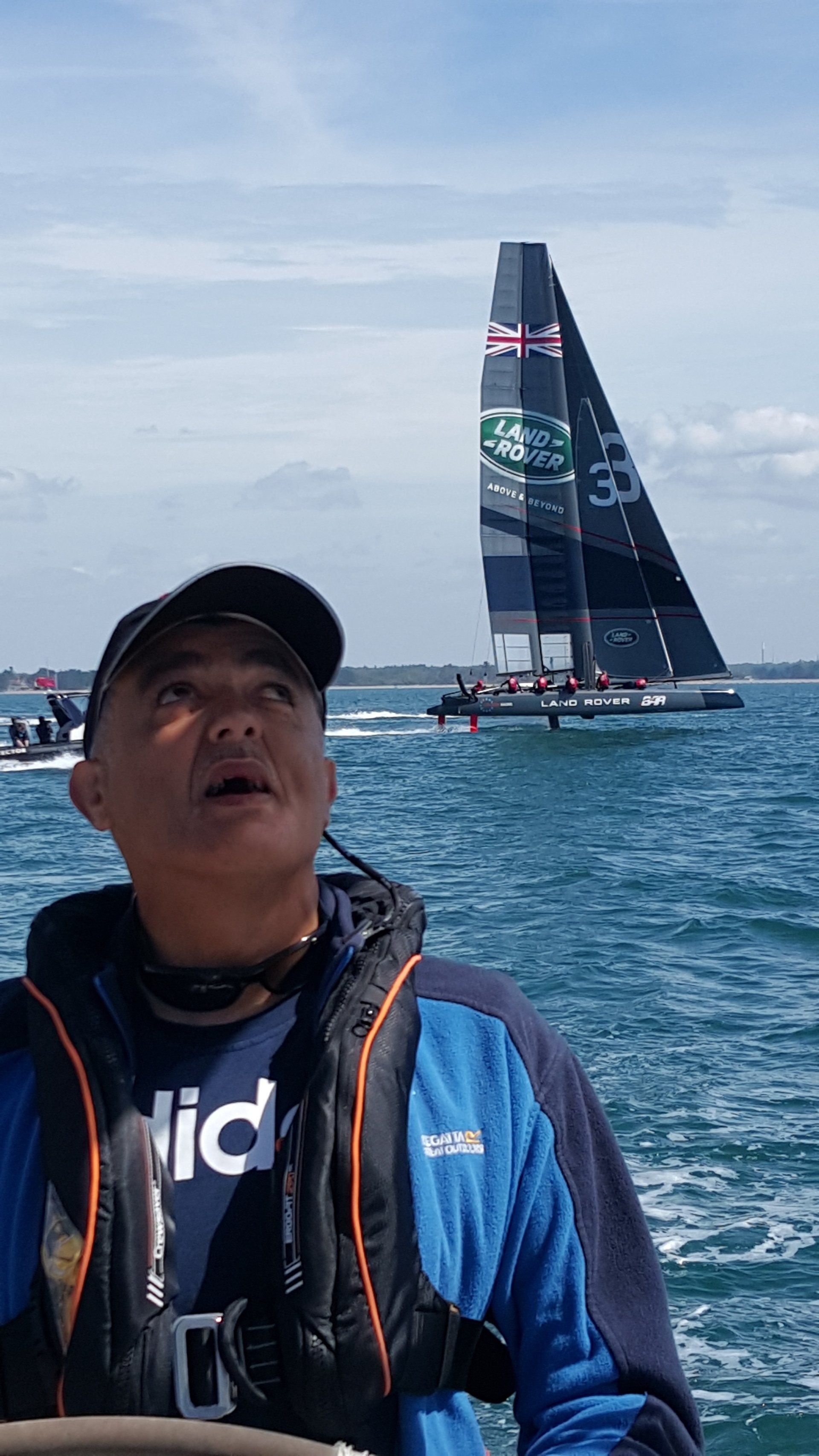 A close up of a V.I. sailor at the helm of an underway yacht. In the background, a yacht of the Land Rover BAR team, skippered by Ben Ainslie, is passing at high speed raised on hydrofoils.
