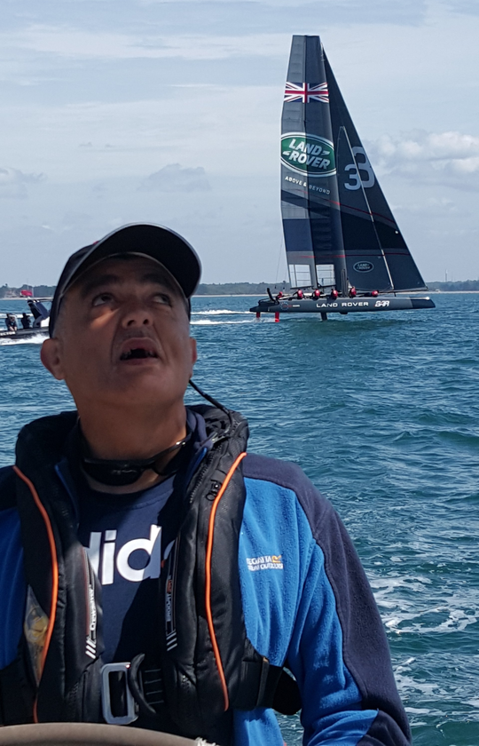 A close up picture of a VI male sailor at the wheel of an underway yacht.  In the background is a racing yacht of the Land Rover BAR team, skippered by Ben Ainslie, passing at high speed raised on aerofoils.