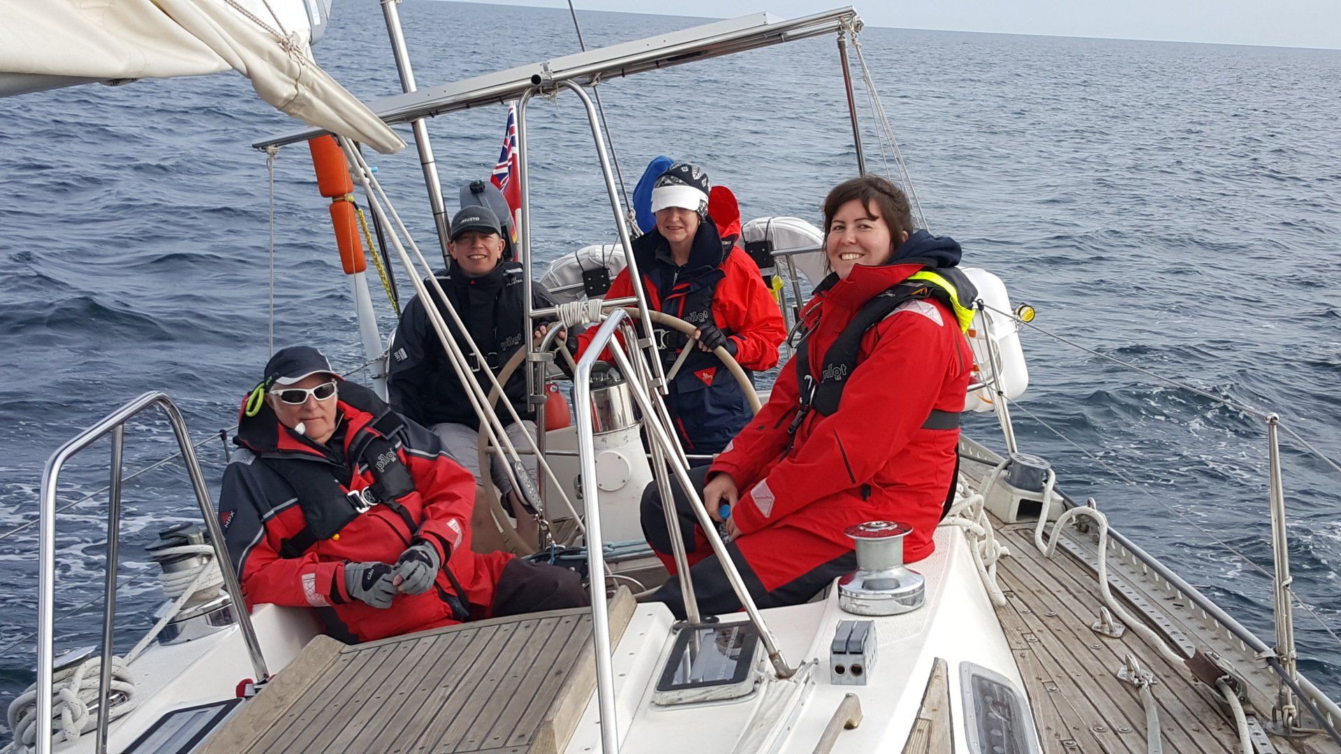 A picture taken from the deck of an underway yacht looking back to the helm. The sighted and V.I. crew are smiling at the camera, dressed in wet weather gear.