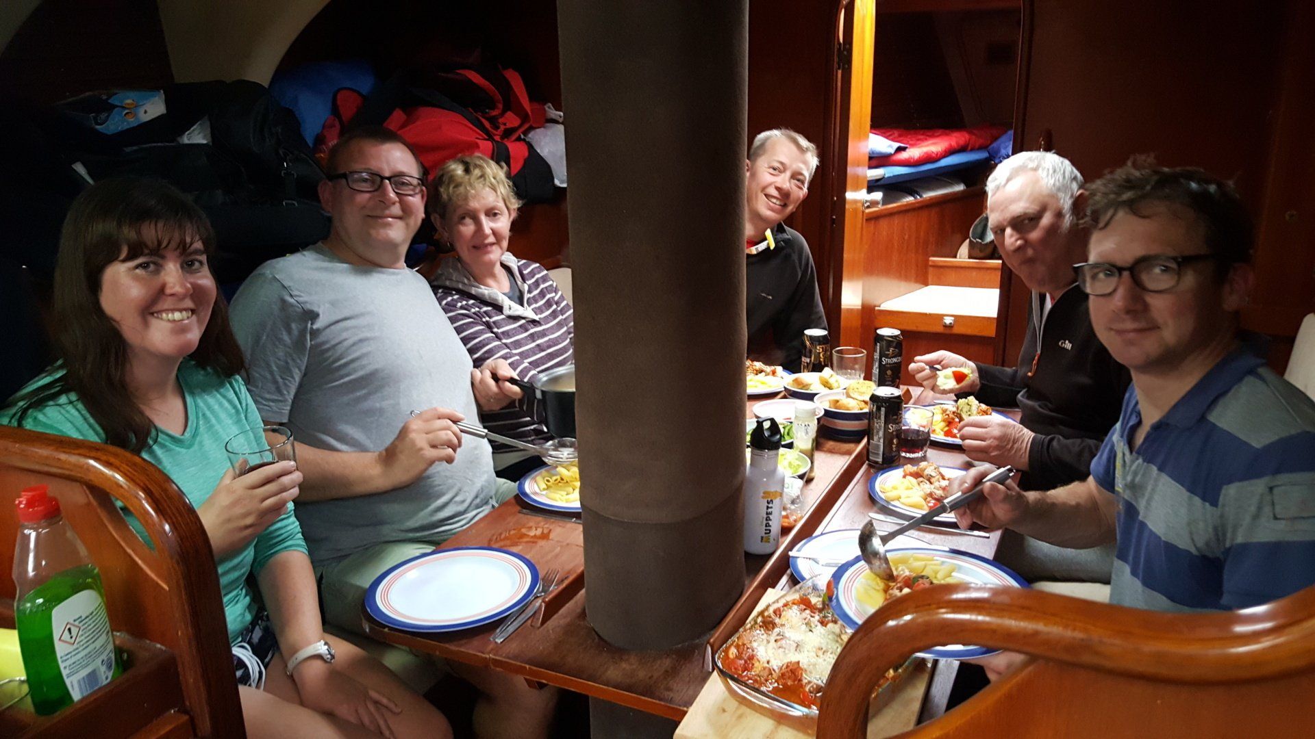 A group of sighted and visually-impaired sailors are sitting around the table below decks, which is set with a meal of pasta, breads and sauces. Everyone is smiling and having fun. The man nearest to the camera is serving some of the food.
