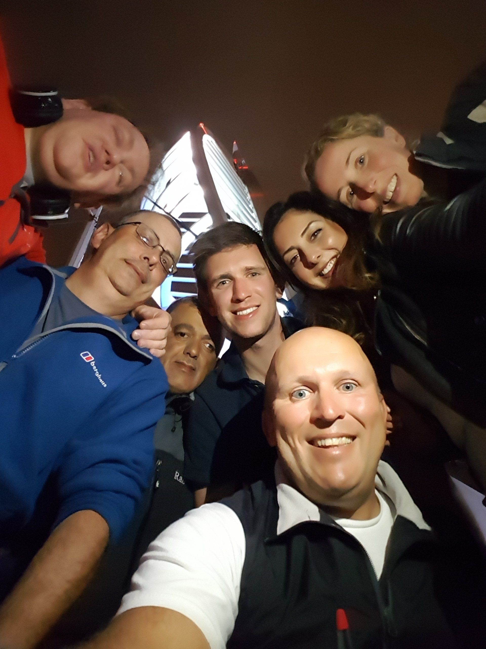 A selfie image of a mixed crew, taken at night from a low angle looking up. Everyone is smiling and the brightly lit Spinnaker Tower in Portsmouth is in the background