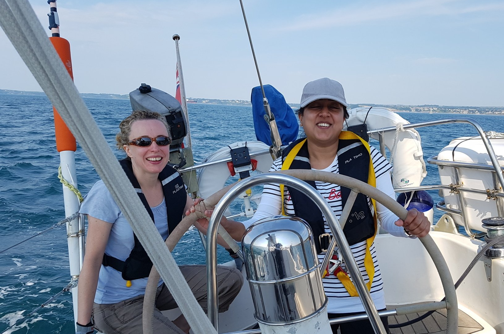 A picture of a smiling female VI sailor at the wheel, and a smiling female sighted crew member next to her. Calm conditions.