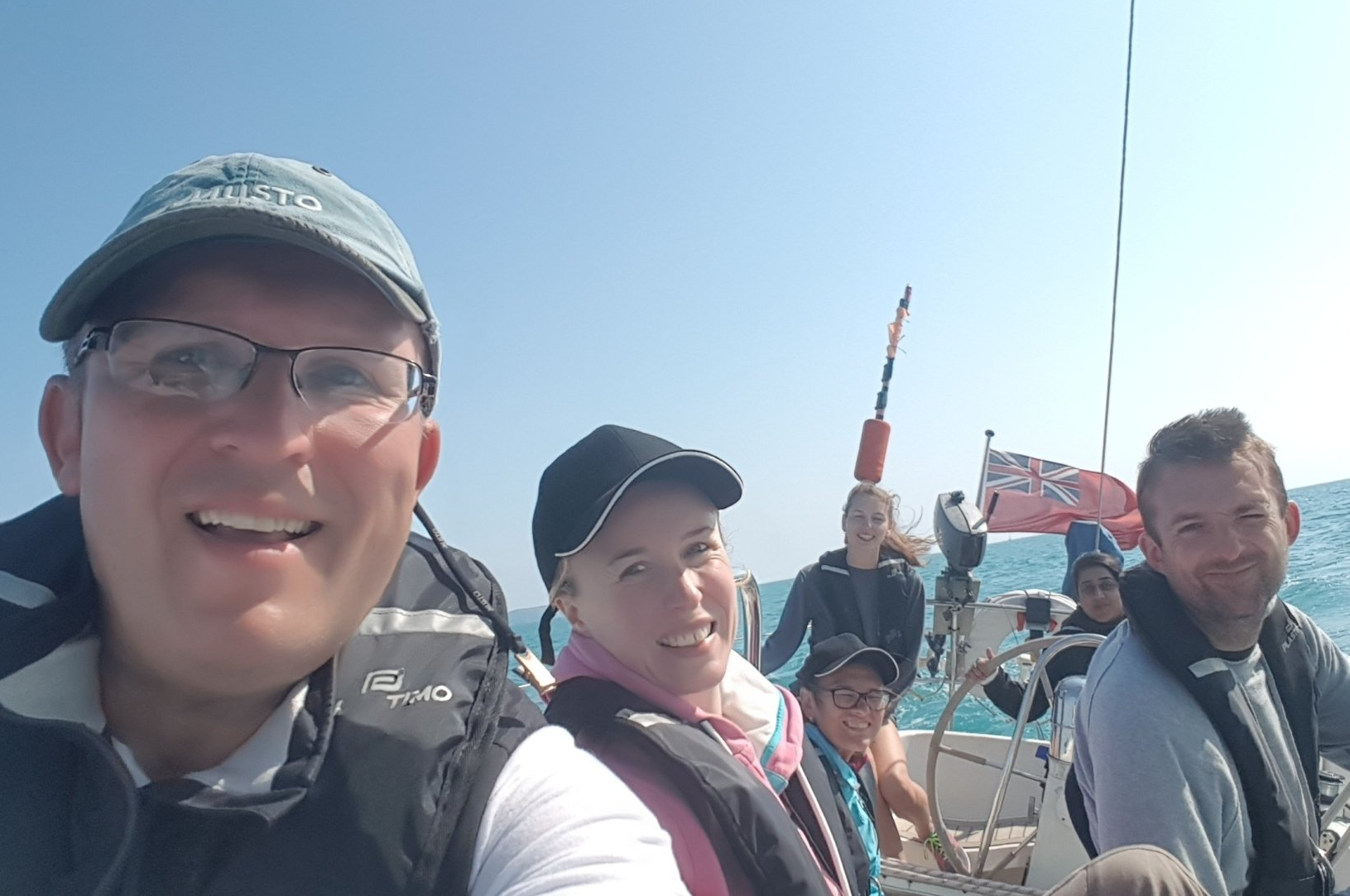 A selfie picture of a smiling crew on an underway yacht. The crew is a mixture of sighted and VI sailors. The horizon in the background is at an angle, showing the yacht is under sail.