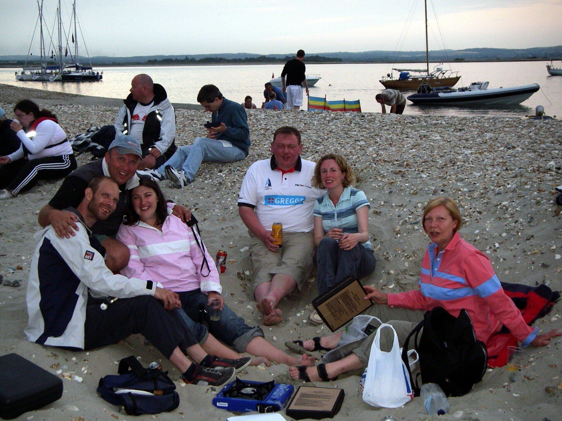 A group of sighted and visually-impaired sailors are sitting on a beach in the evening after a day's sailing. They've got food and drinks and are smiling at the camera. A group has their arms around each others' shoulders. The ocean and moored boats are in the background.