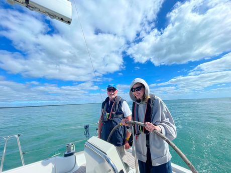 A picture of a sailing yacht underway, taken from the deck looking towards the stern. There is a VI crew member at the wheel with their skipper stood next to them both smiling