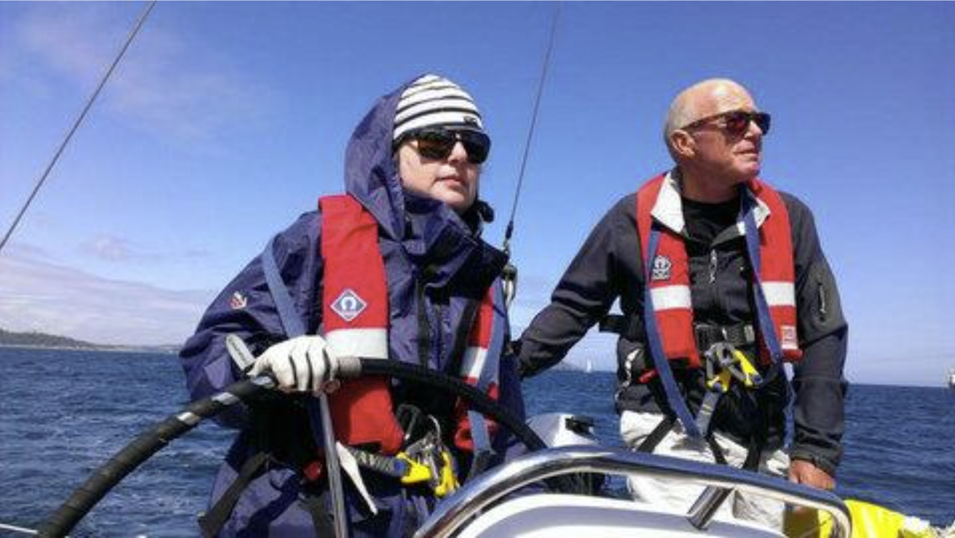 A close up picture of a very focused V.I. sailor at the helm of an underway yacht, with a sighted skipper standing nearby. both are looking towards the bow. The sky is clear and blue, though both are wearing wet weather appropriate gear.